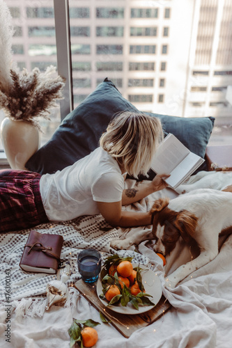 Woman and dog cuddle on bed. Winter cozy vibes. Mandarins, lights, books. Cute puppy. Reading book. Merry Christmas time. Holiday festive season. Cold winters. Staying at home.Warm blanket and pillows