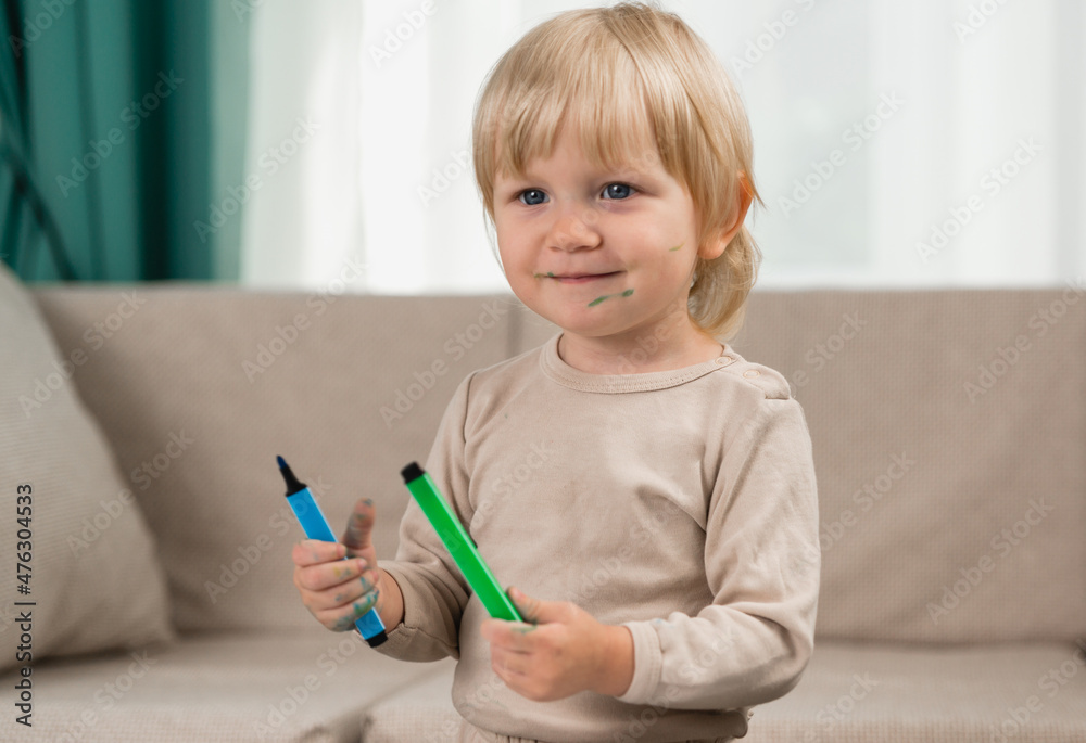 A little cheerful girl in pajamas at home with two felt-tip pens her hands. Face and clothes are stained with paint A little cheerful girl in pajamas at home with two felt-tip pens her hands. Face and clothes are stained with paint