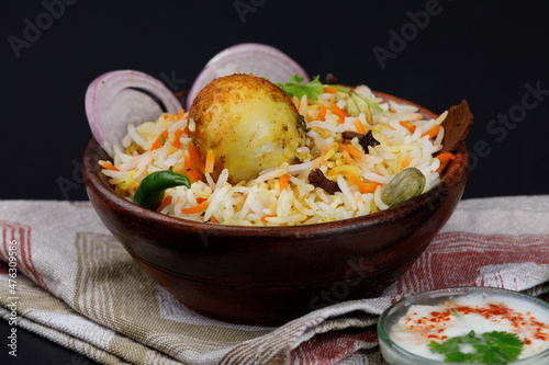 Hot and spicy egg biryani with fried egg served in a wooden bowl with Raita or curd or yogurt on a table napkin isolated on a black background photo