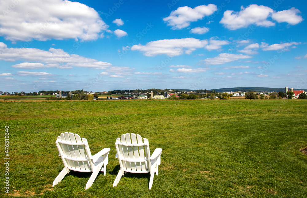 Two empty white Adirondack chairs on a field of green grass overlooking a small town and blue sky with white clouds. Two empty white Adirondack chairs on a field of green grass overlooking a small town and blue sky with white clouds.