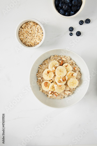 Bowl of porridge topped with banana and almonds,  a bowl of oats and a  bowl of blueberries on white marble countertop