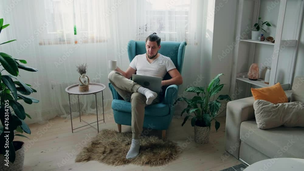 Young man using laptop typing and working sitting on the armchair at home and drink coffee Young man using laptop typing and working sitting on the armchair at home and drink coffee