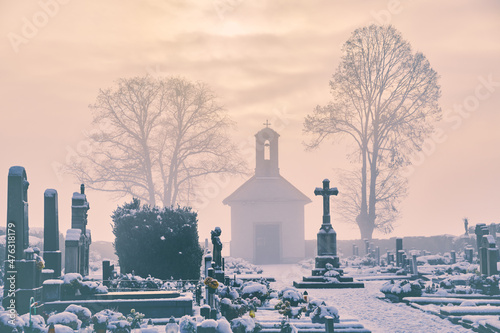 Winter graveyard at christmas with sunny clouds over the cross and morgue. Snowy and foggy cemetery with two linden trees and soft fog above graves with gravestones.