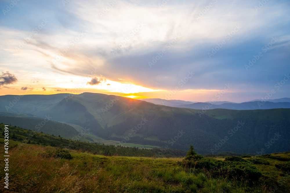 Obraz premium Summer evening mountain landscape with grassy hills and distant peaks at colorful sunset.