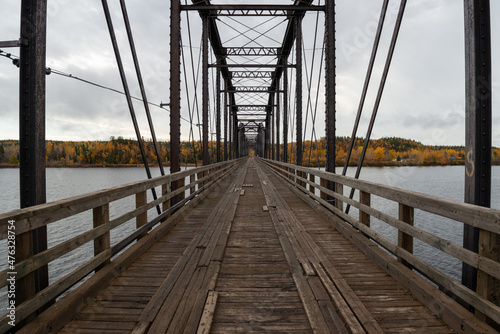 Steel trestle bridge with wooden deck over a large river in Newfoundland. The bridge is for foot traffic and ATV usage. The sky is clear blue and land and houses can be seen in the background. 