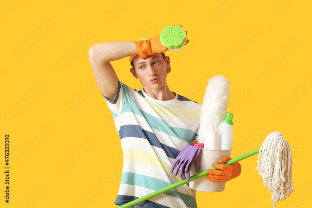 Tired young man with cleaning supplies on yellow background