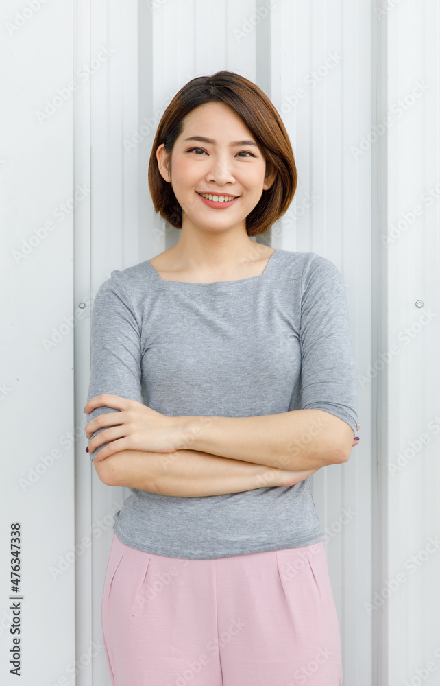 Portrait shot of Asian chinese young happy cheerful millennial short brown hair female model in grey long sleeve shirt and pink pants standing crossed arms smiling look at camera on wall background