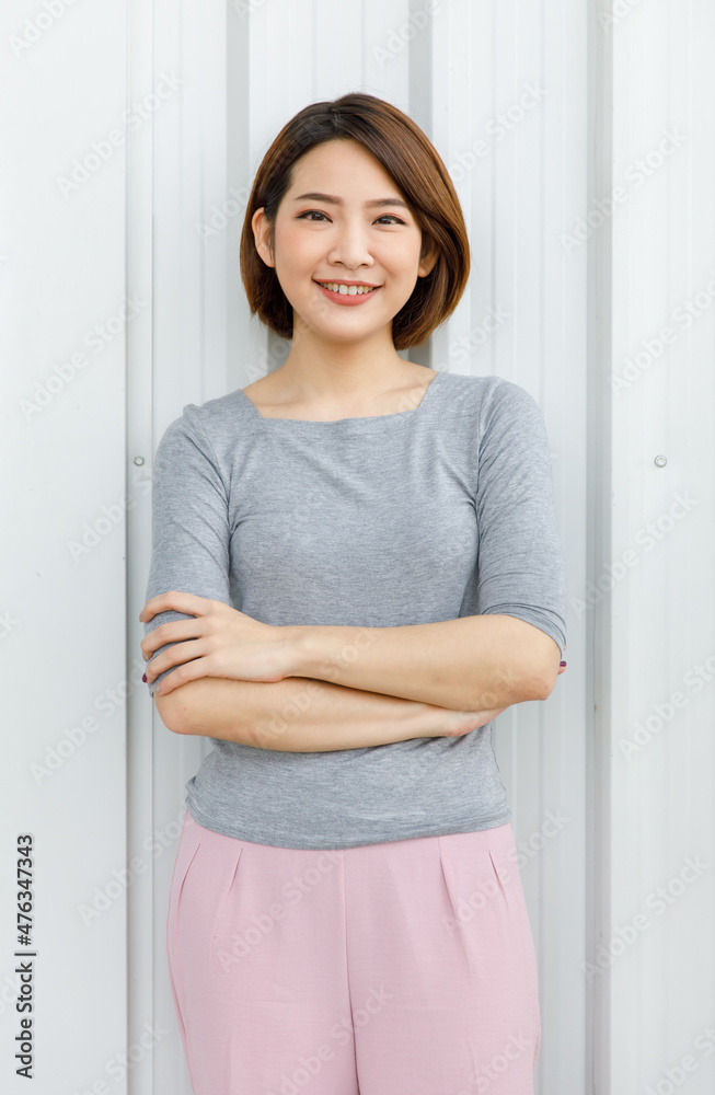 Portrait shot of Asian chinese young happy cheerful millennial short brown hair female model in grey long sleeve shirt and pink pants standing crossed arms smiling look at camera on wall background Portrait shot of Asian chinese young happy cheerful millennial short brown hair female model in grey long sleeve shirt and pink pants standing crossed arms smiling look at camera on wall background
