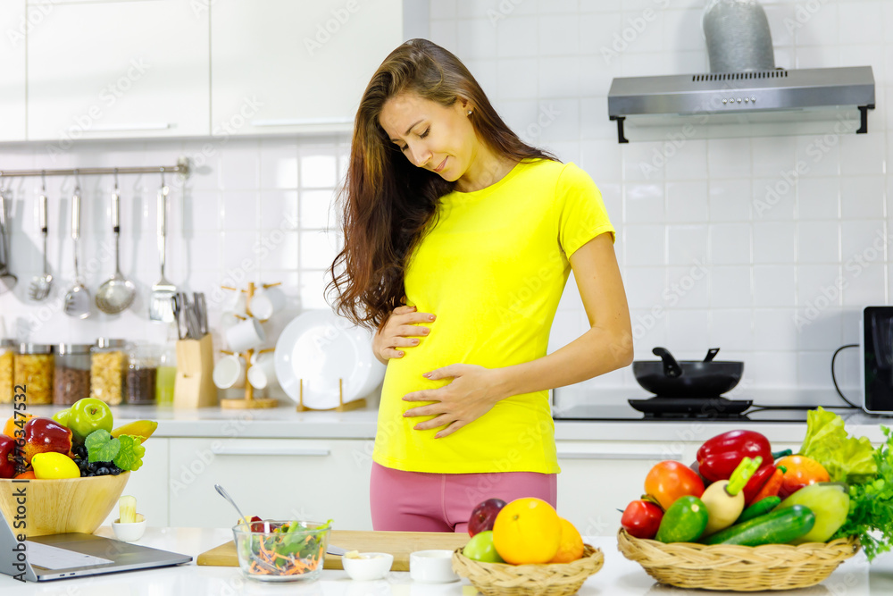 Caucasian young pregnant painful female mother in casual outfit standing holding tummy belly having stomachache while learning to cook vegetable salad online from laptop computer in kitchen at home