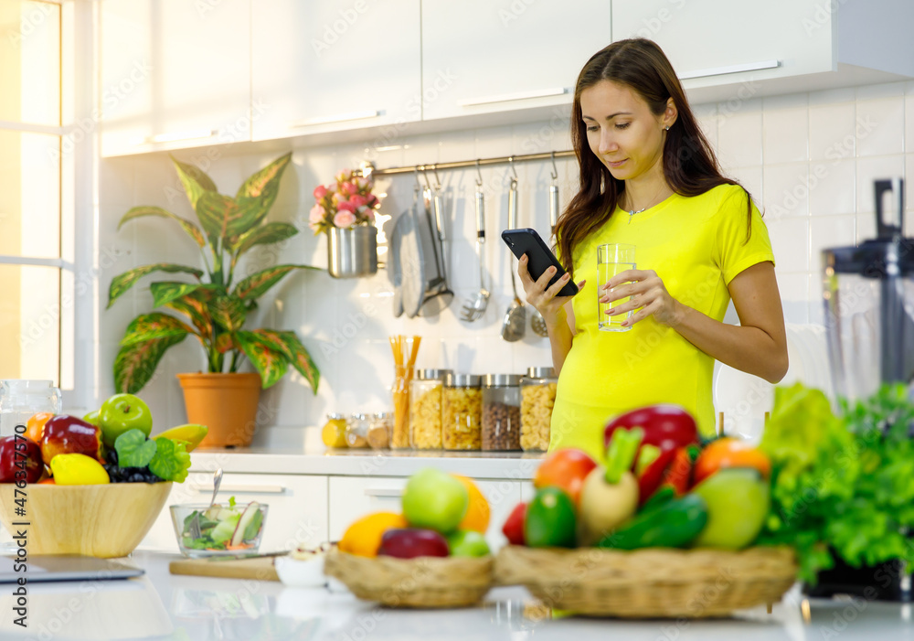 Caucasian young pregnant female mother in casual outfit with big belly tummy stand smiling holding water glass checking email from touchscreen smartphone in kitchen full of cooking equipment at home
