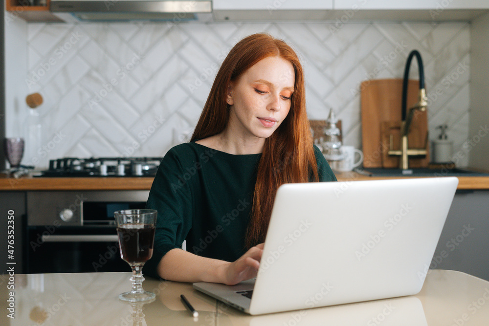 Pretty female freelancer working typing on laptop computer sitting at table with glass of alcoholic beverage in kitchen. Redhead young woman texting e-mail response to client from home office. Pretty female freelancer working typing on laptop computer sitting at table with glass of alcoholic beverage in kitchen. Redhead young woman texting e-mail response to client from home office.
