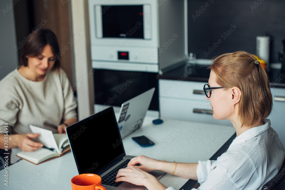Woman's co-working, remotely work, home office, startup. Two attractive female freelancers sitting with laptops face to face in home kitchen. Woman's co-working, remotely work, home office, startup. Two attractive female freelancers sitting with laptops face to face in home kitchen.