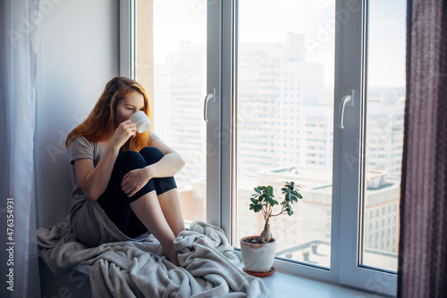 Beautiful young woman sitting on windowsill overlooking the metropolis, drinking tea or coffee from cup. Female portrait on the background of window in city apartment. Peace of mind and mental health.