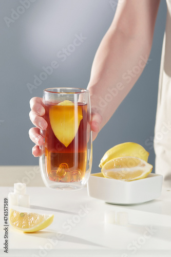 Shot of a female hand holding a glass of black tea with lemon. There are three cubes of refined sugar, a slice of lemon and lemons on the table. Items on the gray background.