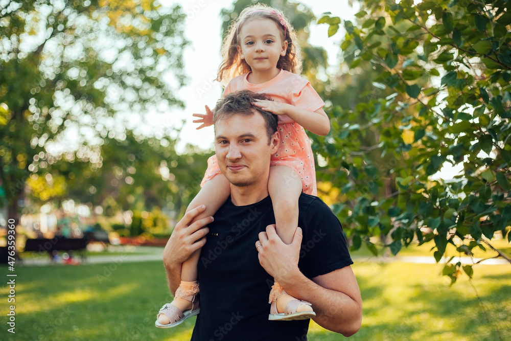 a girl is sitting on dad's shoulders on a walk in a summer park 
