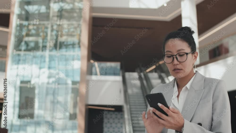 Tracking shot of Asian businesswoman in formal wear and glasses walking through office building lobby and typing on mobile phone Tracking shot of Asian businesswoman in formal wear and glasses walking through office building lobby and typing on mobile phone