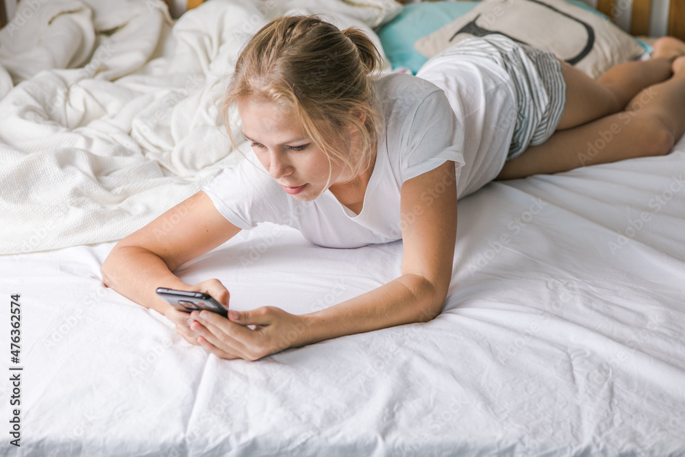 Online dating. Romantic. Portrait of a smiling middle aged girl lying on bed with mobile phone. Happy blonde woman using cellphone at home. Beautiful young lady typing on smartphone in her bedroom. Online dating. Romantic. Portrait of a smiling middle aged girl lying on bed with mobile phone. Happy blonde woman using cellphone at home. Beautiful young lady typing on smartphone in her bedroom.