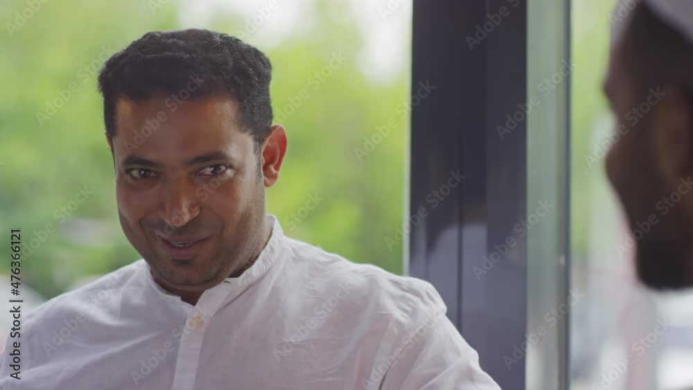 Shoulder-up of young Middle Eastern man wearing white shirt sitting in cafe by window at daytime, talking to cropped friend, smiling Shoulder-up of young Middle Eastern man wearing white shirt sitting in cafe by window at daytime, talking to cropped friend, smiling