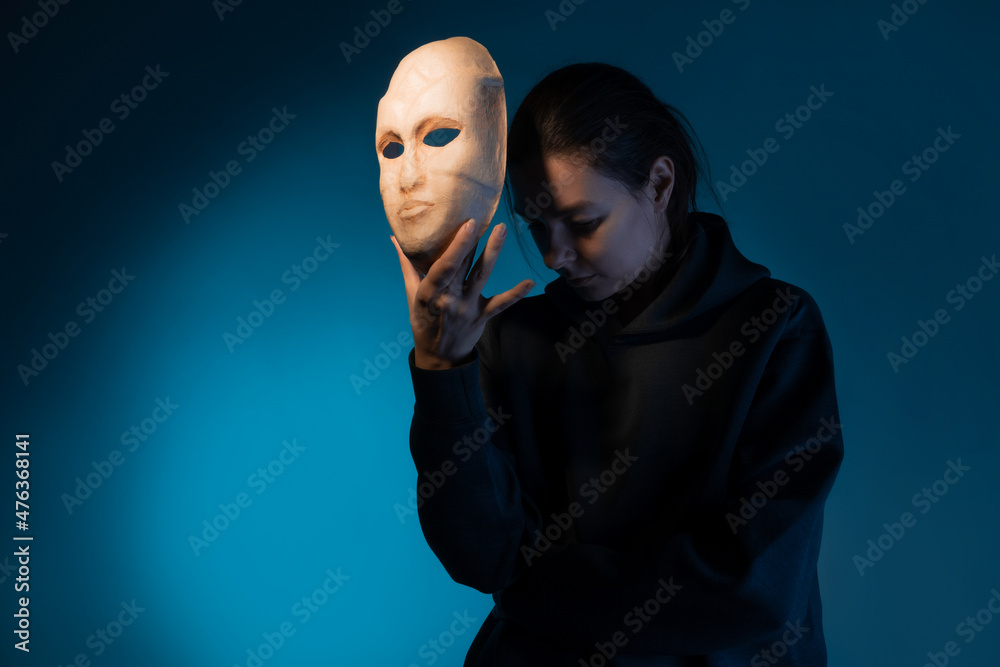 Hiding behind a mask, a young woman in a dark hoodie hides her face with a mask, self-identification problems and impostor syndrome. Portrait in the studio on a dark gray background. - obrazy, fototapety, plakaty 
