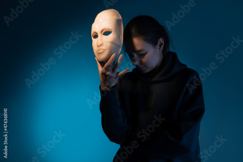 фотография Hiding behind a mask, a young woman in a dark hoodie hides her face with a mask, self-identification problems and impostor syndrome