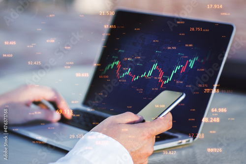 Close up of businessman hands using laptop and smartphone with abstract candlestick forex graph and bid data numbers on blurry desktop background. Finance, technology, analytics and trade concept. 