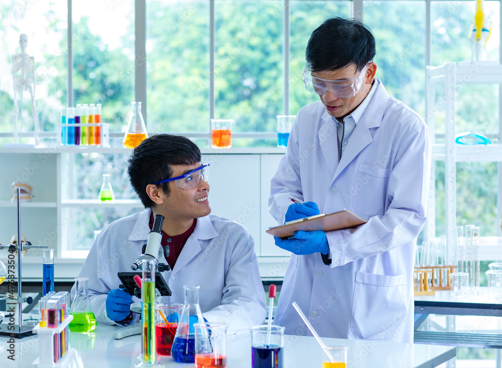 Asian young university male scientist student in white lab coat safety goggles and rubber gloves using microscope magnifying science sample in laboratory while teacher holding clipboard rating score Asian young university male scientist student in white lab coat safety goggles and rubber gloves using microscope magnifying science sample in laboratory while teacher holding clipboard rating score
