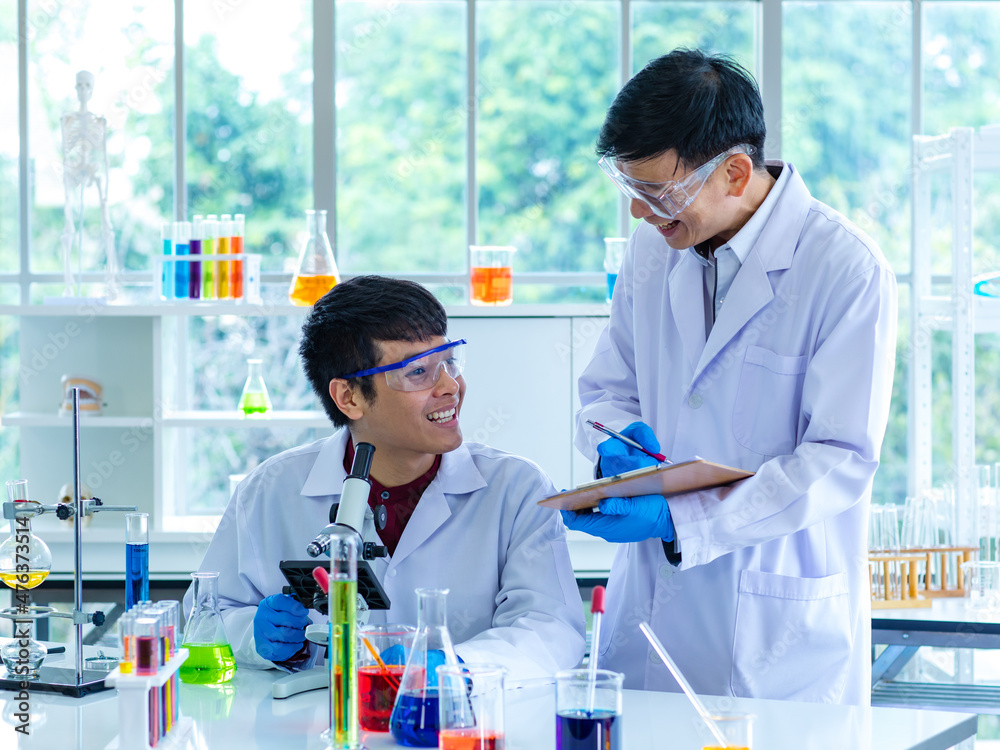 Asian young university male scientist student in white lab coat safety goggles and rubber gloves using microscope magnifying science sample in laboratory while teacher holding clipboard rating score Asian young university male scientist student in white lab coat safety goggles and rubber gloves using microscope magnifying science sample in laboratory while teacher holding clipboard rating score
