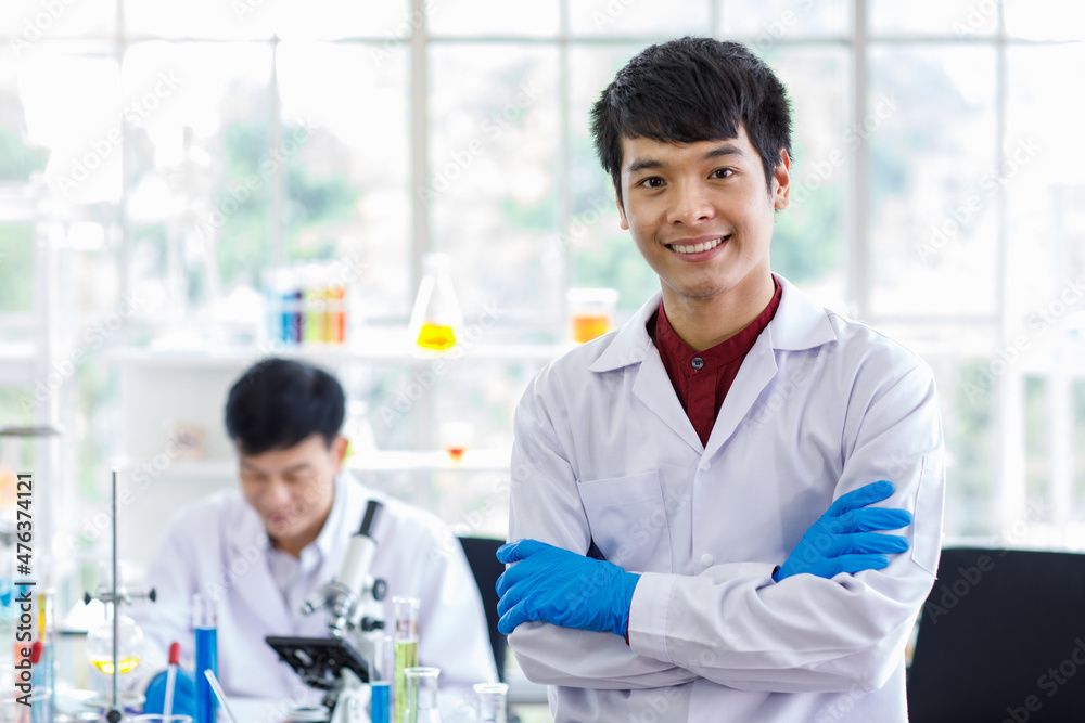 Portrait shot of Asian professional successful young handsome male scientist in white lab coat rubber gloves standing crossed arms smiling look at camera in laboratory while colleague working behind Portrait shot of Asian professional successful young handsome male scientist in white lab coat rubber gloves standing crossed arms smiling look at camera in laboratory while colleague working behind