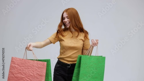 . A teenage girl in an orange T-shirt with red hair, laughs and dances with colorful shopping bags in her hands. Posing on a gray background.