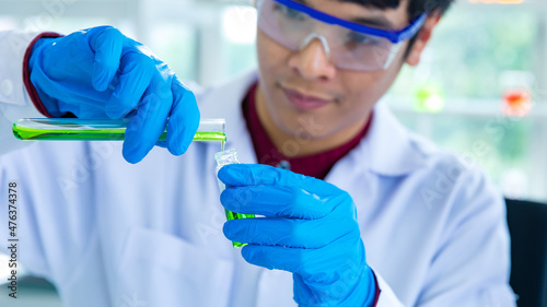 Closeup shot hands of Asian male scientist in white lab coat safety goggles and rubber gloves in blurred background careful holding pouring green sample from test tube to glass vial in laboratory