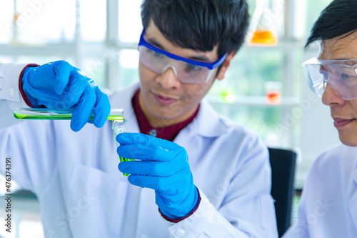 Closeup shot hands of Asian male scientist in white lab coat safety goggles and rubber gloves in blurred background careful holding pouring green sample from test tube to glass vial in laboratory photo