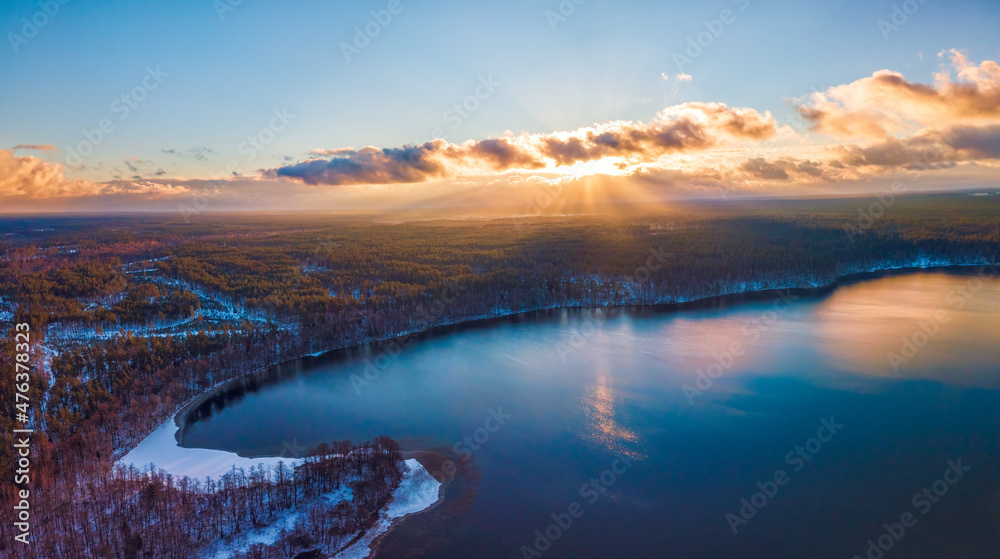 Aerial view of the winter background with a snow-covered forest, lake and sunrays over the Glukas lake in Lithuania