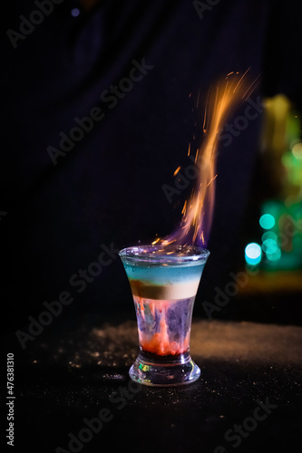 The fire scatters into sparks. In the glass, the cocktail is brown-red the top is white and the top is emerald which is set on fire, layers are not mixed Filming of the bar menu against the background