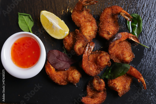 Shrimp is served with sauce. Tempura king prawns, sliced lemon, green basil leaves are served on a black stone stand. Cutlery, hot pepper pods, spices in bowls, white mushrooms.