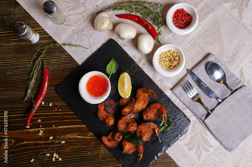 Shrimp is served with sauce. Tempura king prawns, sliced lemon, green basil leaves are served on a black stone stand. Cutlery, hot pepper pods, spices in bowls, white mushrooms.