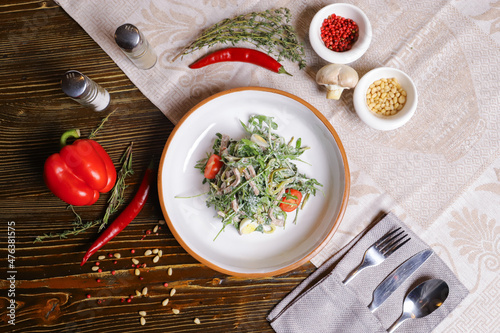 On a dark countertop, a light linen napkin, on a cutting board Served salad with rucola leaves, boiled tongue cut into long slices with sauce A couple of salt and pepper, cutlery, red peppers decorate