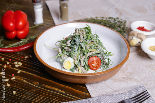 On a dark countertop, a light linen napkin, on a cutting board Served salad with rucola leaves, boiled tongue cut into long slices with sauce A couple of salt and pepper, cutlery, red peppers decorate