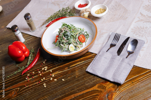 On a dark countertop, a light linen napkin, on a cutting board Served salad with rucola leaves, boiled tongue cut into long slices with sauce A couple of salt and pepper, cutlery, red peppers decorate