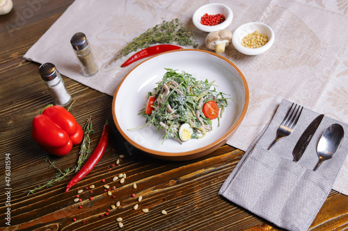 On a dark countertop, a light linen napkin, on a cutting board Served salad with rucola leaves, boiled tongue cut into long slices with sauce A couple of salt and pepper, cutlery, red peppers decorate
