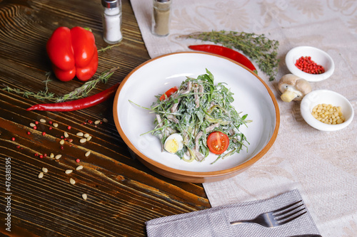 On a dark countertop, a light linen napkin, on a cutting board Served salad with rucola leaves, boiled tongue cut into long slices with sauce A couple of salt and pepper, cutlery, red peppers decorate