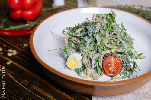 On a dark countertop, a light linen napkin, on a cutting board Served salad with rucola leaves, boiled tongue cut into long slices with sauce A couple of salt and pepper, cutlery, red peppers decorate