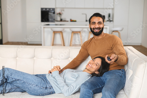 Horizontal shot of the Indian couple in love sitting at the sofa and watching TV with pleasure smiles. Multiracial man holding remote controller at hand. Home concept © Vadim Pastuh