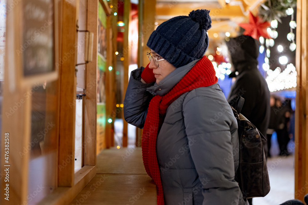 caucasian woman wearing hat and red scarf going to buy buying treats in kiosk at christmas market