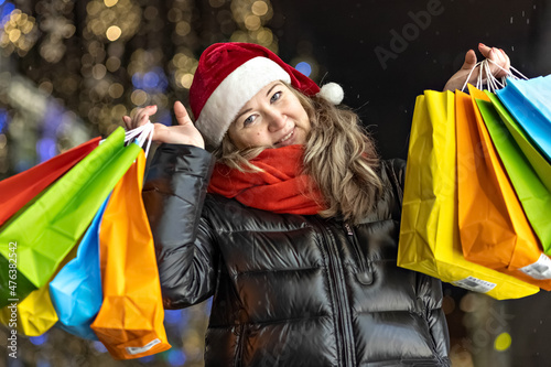 Portrait of a woman in a Santa hat with Christmas shopping in colorful bags on the street decorated with Christmas lights and decorations © Светлана Густова