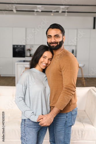 Smiling man hugging his charming woman. Two people standing and joyfully looking at the camera. Young Indian couple happily spending time in cozy modern kitchen at home