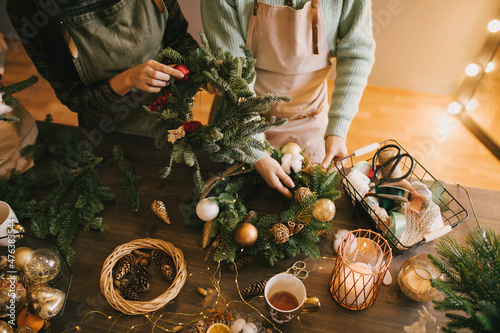 Two millennial women making Christmas wreath using pine branches and festive decorations. Small business photo