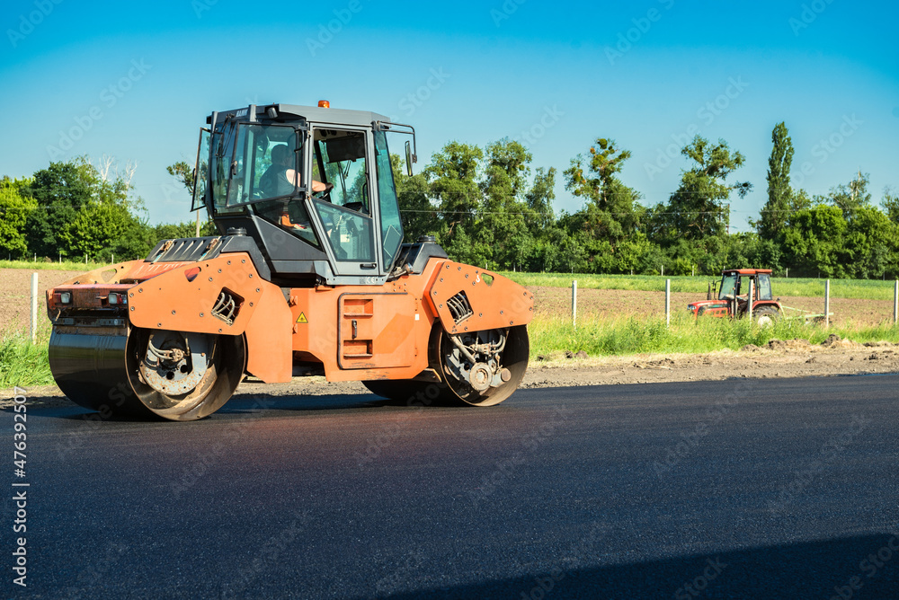 Rollers rolling fresh hot asphalt on the new road with a plowing tractor in the background. Road construction. Rollers rolling fresh hot asphalt on the new road with a plowing tractor in the background. Road construction.