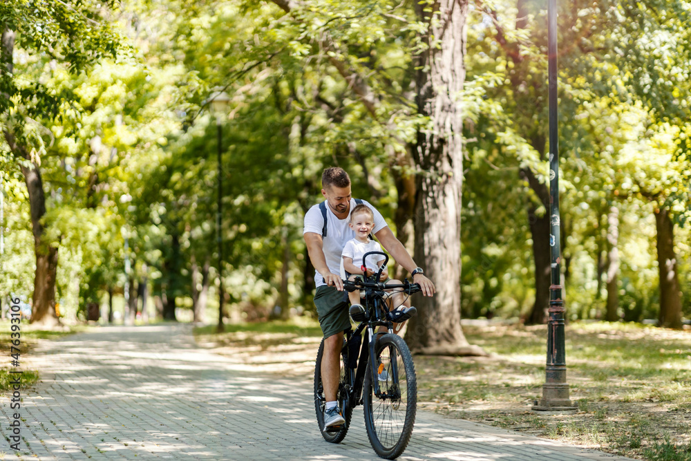 Active family vacation and match casual clothes. A male toddler and a father ride a bike around the park. Cycling holiday and active weekend. Toddler sits in the basket on the bike and laugh with dad