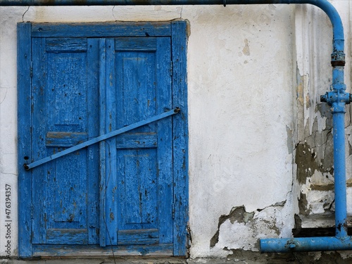 a fragment of the facade of an old destroyed building with white fallen off plaster and blue shutters of a closed window and a peeling blue gas pipe, a dilapidated abandoned building outside view