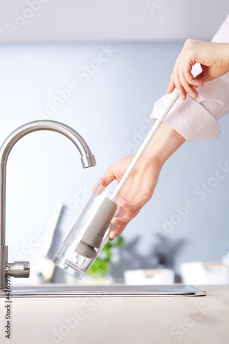 Subject shot of a person washing a glass with a special sponge. The sponge has a long white handle. The glass and sponge are under the tap. Kitchen items are on the gray background.